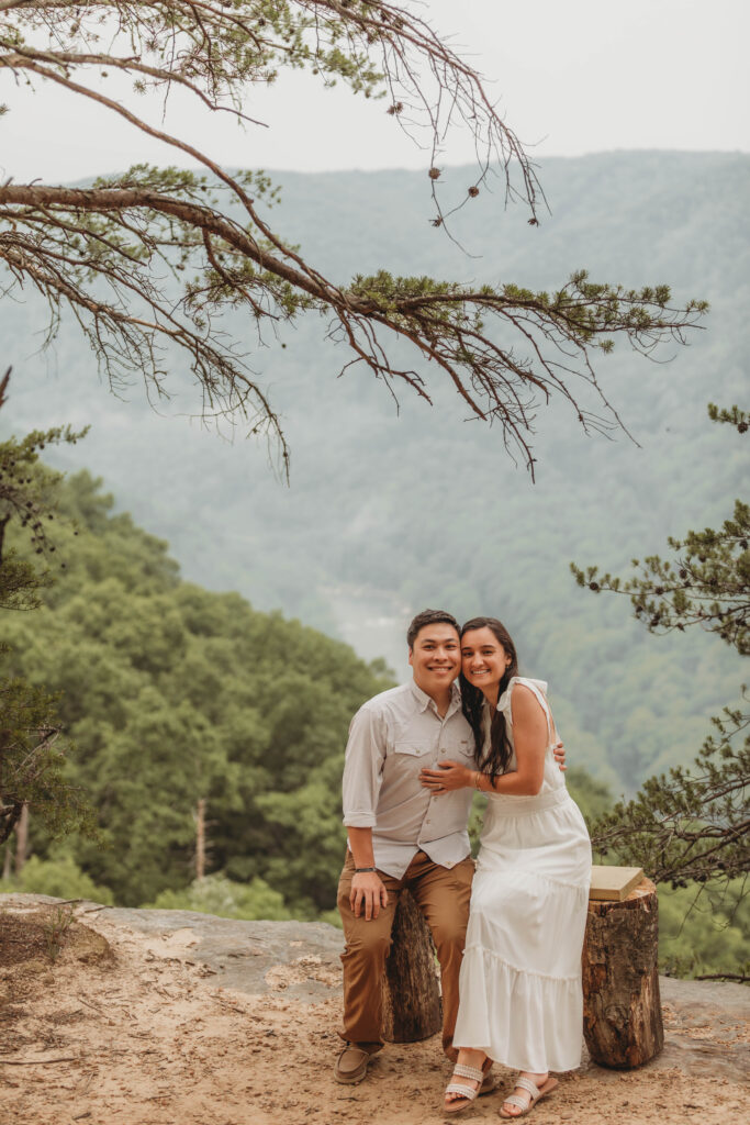 A couple sit on a wooden stump and smile at the camera during their engagement photos at the New River Gorge.