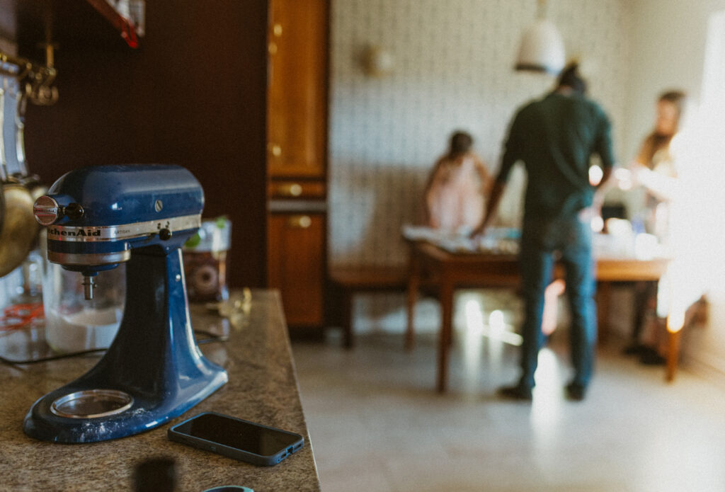 A phone is seen laying on a table next to a kitchen mixer with a family in the background cooking together during in home documentary photos.